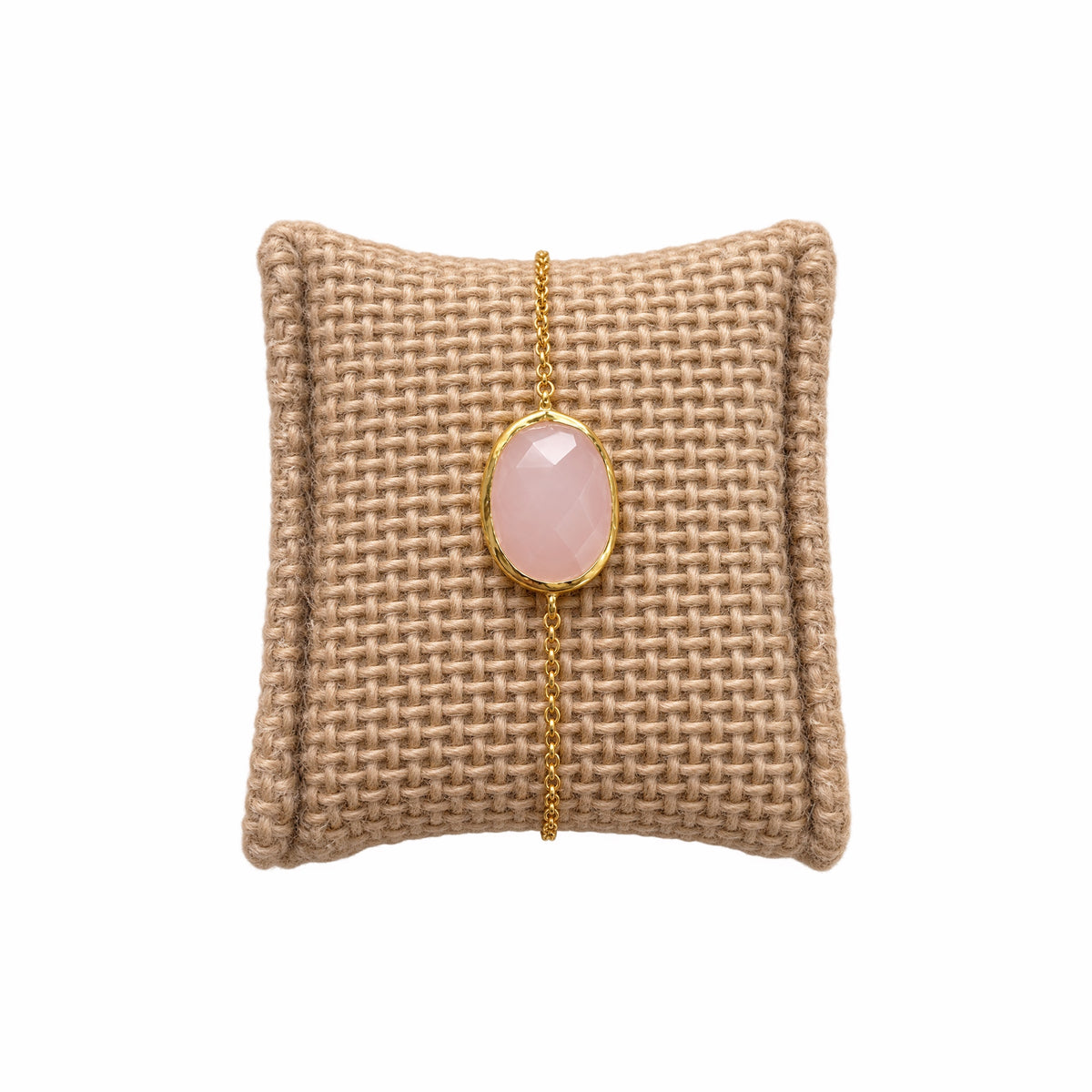 A minimal gold chain bracelet featuring an oval faceted pink rose quartz gemstone, on a textured beige woven cushion against a solid white background.