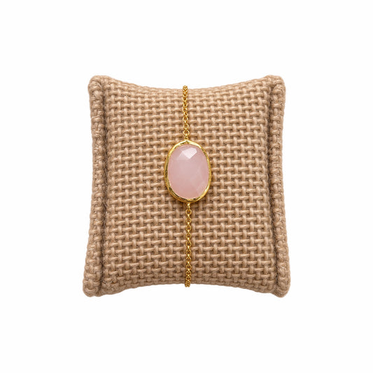 A minimal gold chain bracelet featuring an oval faceted pink rose quartz gemstone, on a textured beige woven cushion against a solid white background.