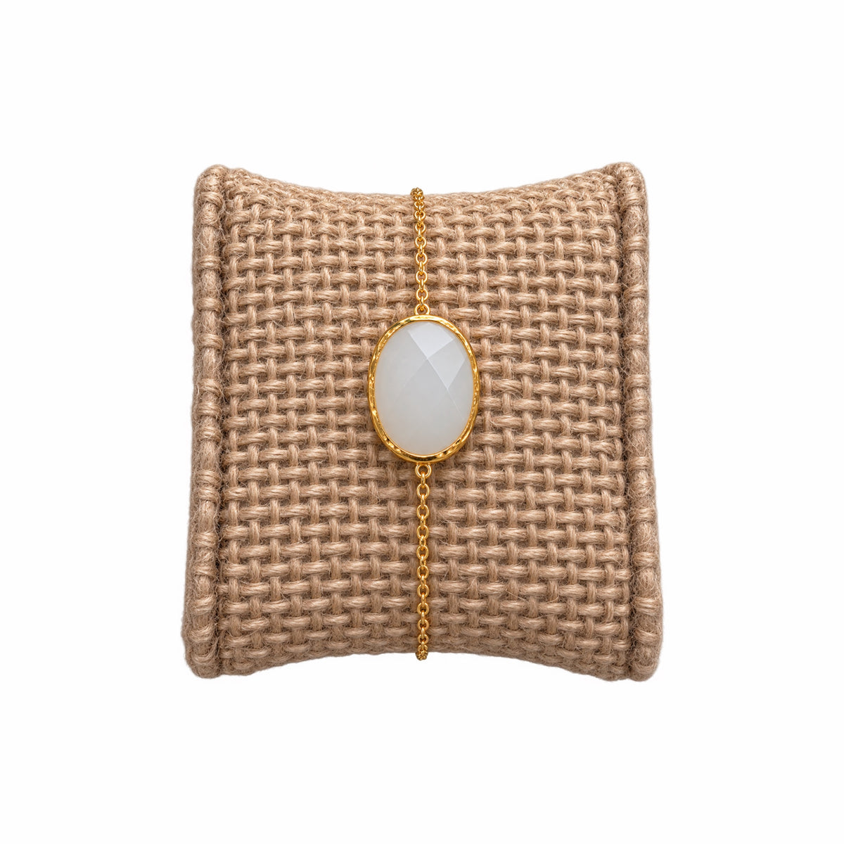 A minimal gold chain bracelet with oval faceted white snow Quartz gemstone, on a woven beige jewelry cushion against a white background.