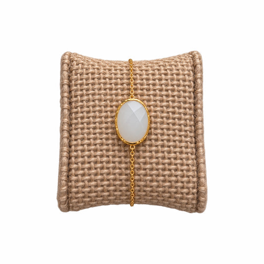A minimal gold chain bracelet with oval faceted white snow Quartz gemstone, on a woven beige jewelry cushion against a white background.