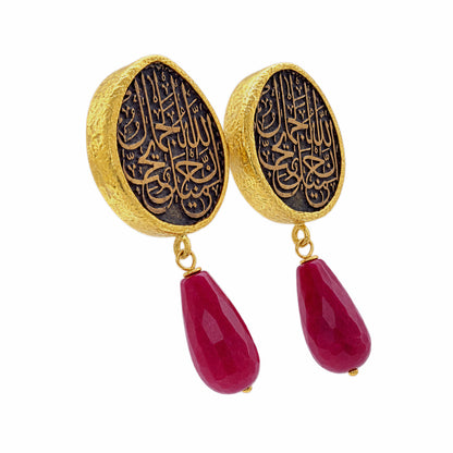 A pair of gold and black drop earrings featuring circular studs with Arabic calligraphy and dangling red ruby on a white background
Facing right