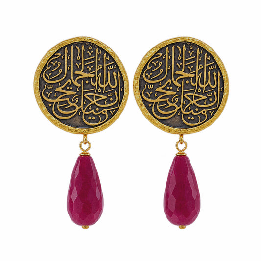 A pair of gold and black drop earrings featuring circular studs with Arabic calligraphy and dangling red ruby on a white background
