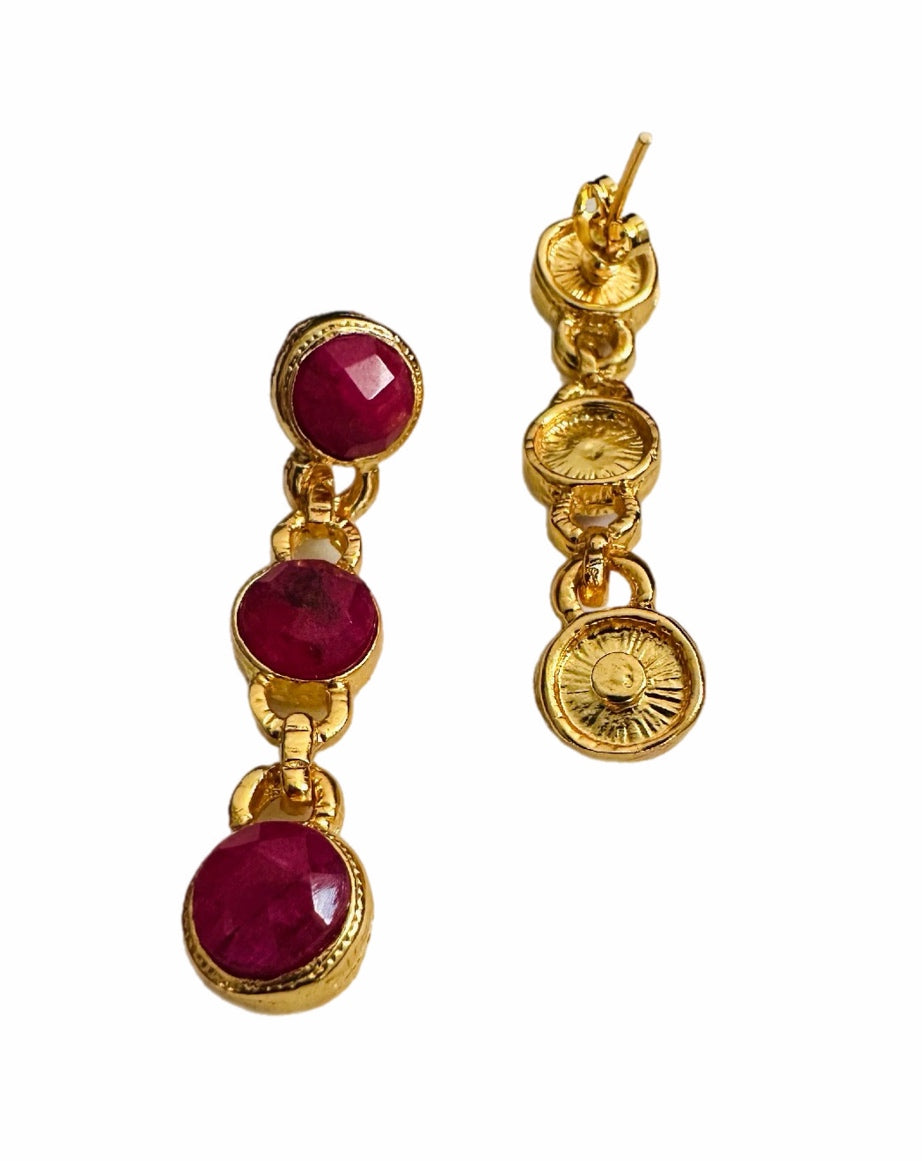 A pair of gold dangle earrings featuring three round, faceted deep red ruby gemstones in each earring, set in gold-toned metal with decorative circular details.