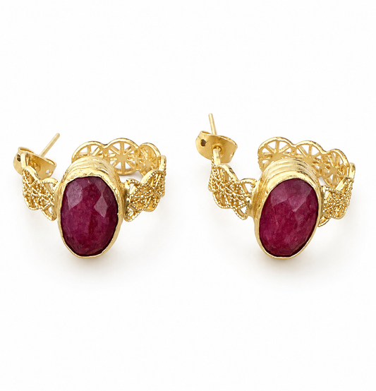 A pair of gold huggie hoop earrings with oval faceted dark red ruby gemstone centers and ornate filigree gold sides, shown on a plain white background.