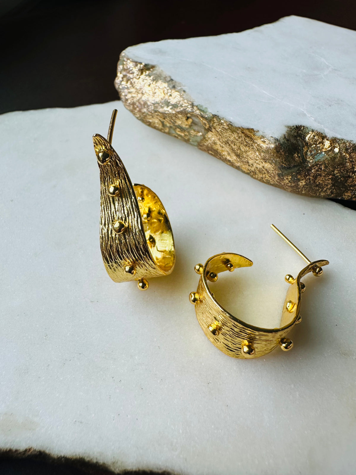 A pair of textured contemporary gold hoop earrings with small raised gold ball accents, on a white marble background.