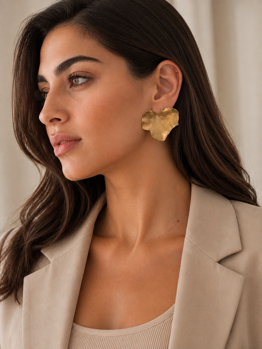 A woman in a beige blazer wearing a textured, leaf-shaped gold statement earrings, shown in a close-up profile portrait against a soft beige background.