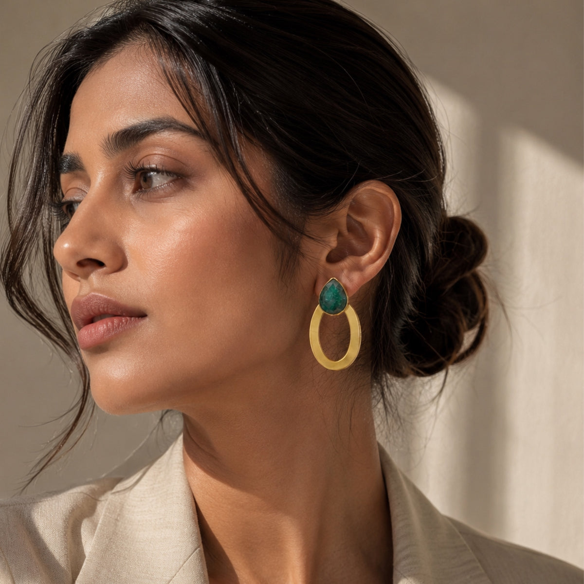 A woman with dark hair in a low bun wearing a gold dangle emerald gemstone earring, shown in a close-up profile against a neutral light gray background.
