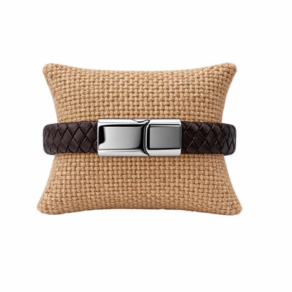 A dark brown woven leather bracelet with polished silver hardware, displayed on a woven tan pillow against a solid white background.