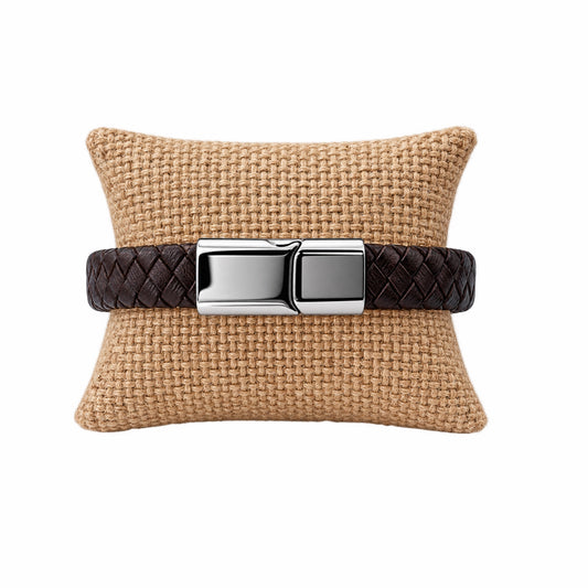 A dark brown woven leather bracelet with polished silver hardware, displayed on a woven tan pillow against a solid white background.