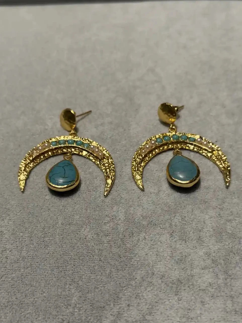 A pair of gold crescent-shaped drop earrings with pearl and turquoise blue stone accents and teardrop-shaped blue stones, displayed on a textured gray surface.