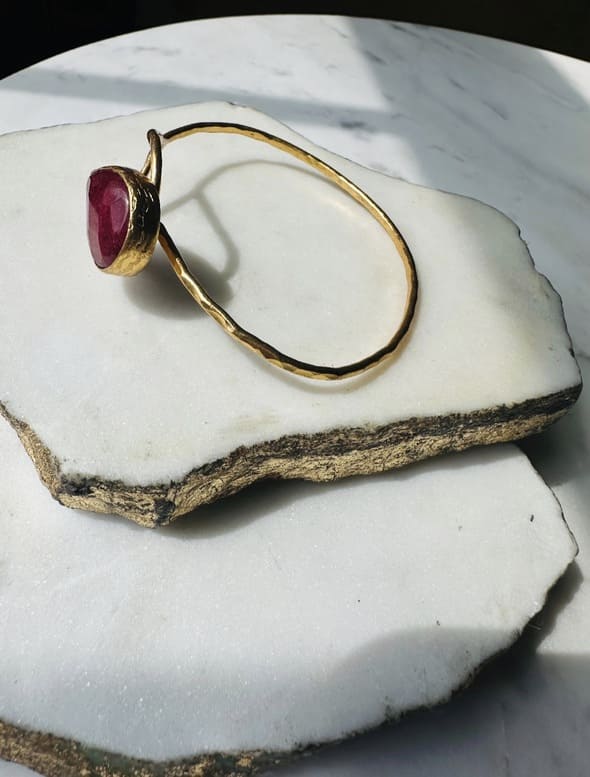 Gold-plated sustainable bangle with a red ruby gemstone on a marble surface