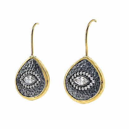 A pair of gold and dark silver teardrop-shaped drop earrings with a hammered gold finish, a textured center, and a central clear zircon evil  eye gemstone surrounded by small clear stones, shown on white background.