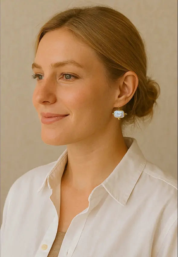 Woman wearing handmade sustainable gold-plated flat pearl rectangular earrings and a white shirt with a neutral background