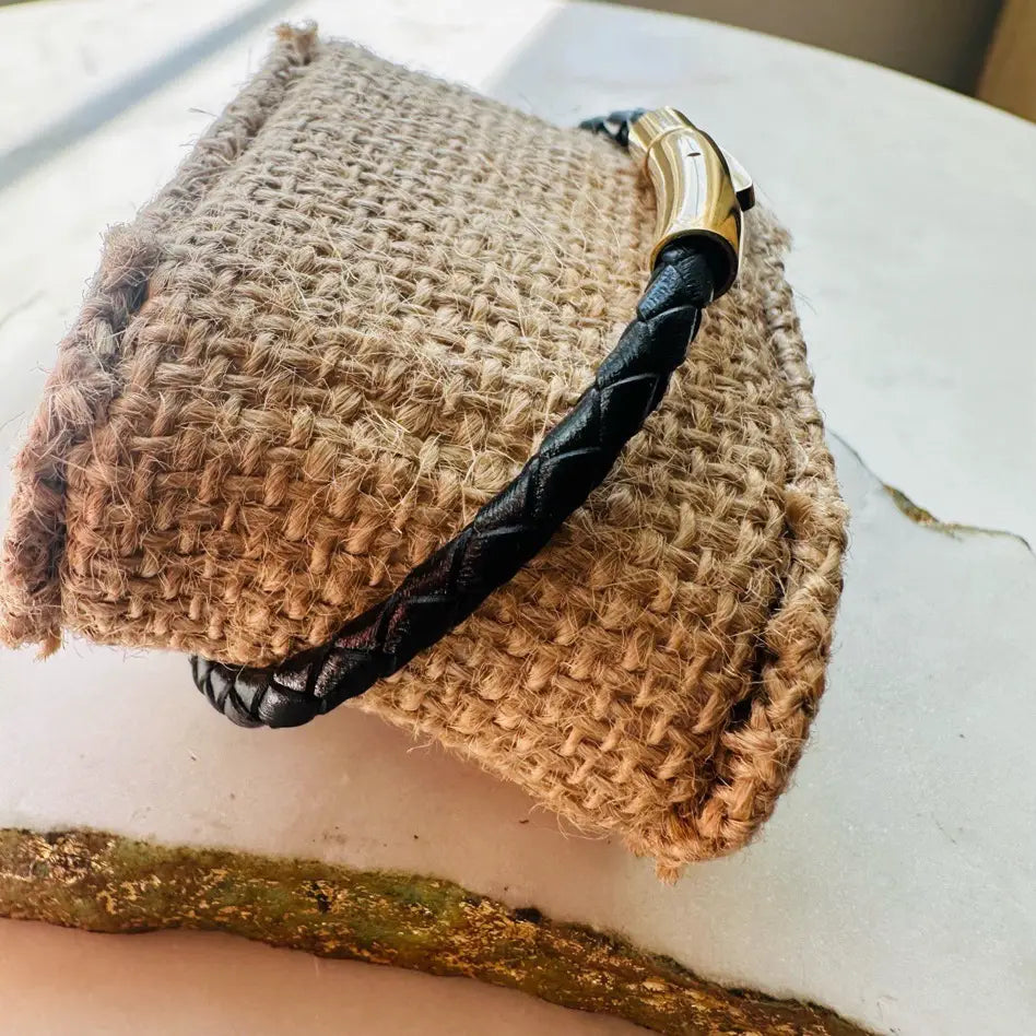 Handmade men’s thin black braided Bracelet with gold clasp details on a textured beige surface