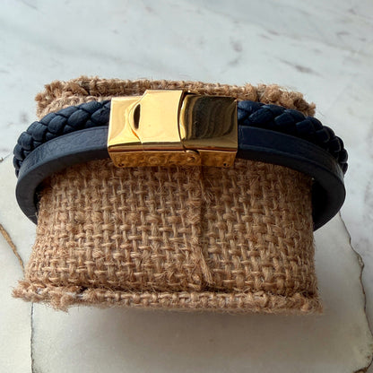 A dark navy blue leather bracelet with a braided front section and a polished gold-tone rectangular clasp, displayed on a textured burlap on marble background.