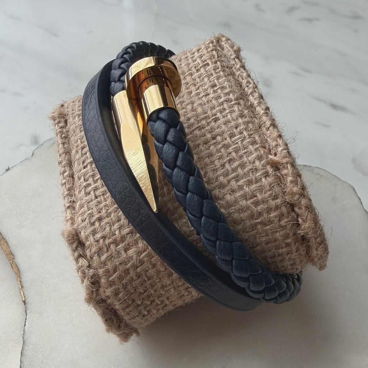 A dark navy blue braided leather wrap bracelet with a polished gold metal chevron clasp, displayed on a textured burlapl.