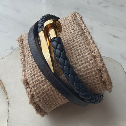 A dark navy blue braided leather wrap bracelet with a polished gold metal chevron clasp, displayed on a textured burlapl.
