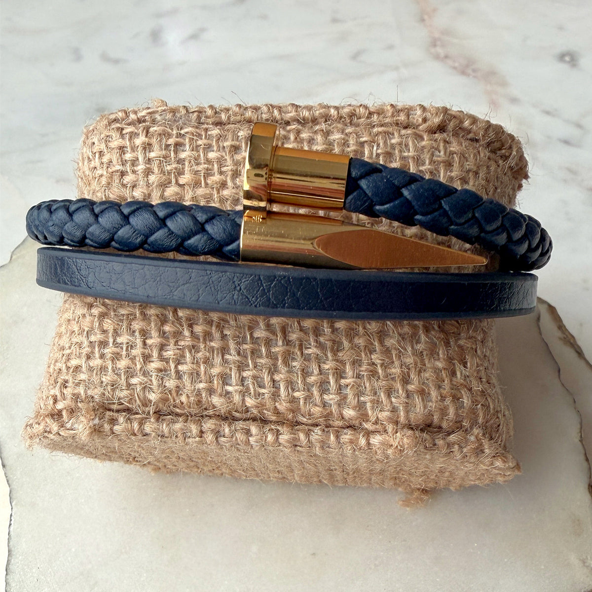 A navy blue leather bracelet set displayed on a burlap ring stand. The set includes a smooth navy leather band with a gold nail-shaped clasp and a braided navy leather bracelet with a matching gold clasp.