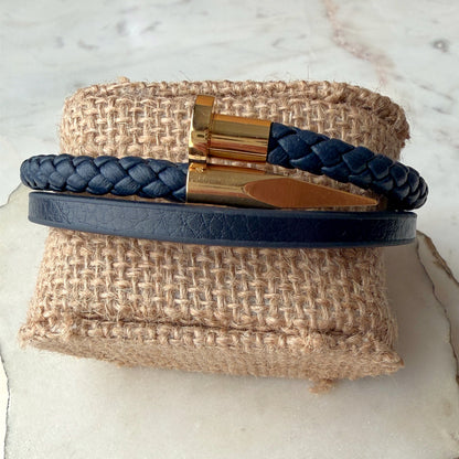 A navy blue leather bracelet set displayed on a burlap ring stand. The set includes a smooth navy leather band with a gold nail-shaped clasp and a braided navy leather bracelet with a matching gold clasp.