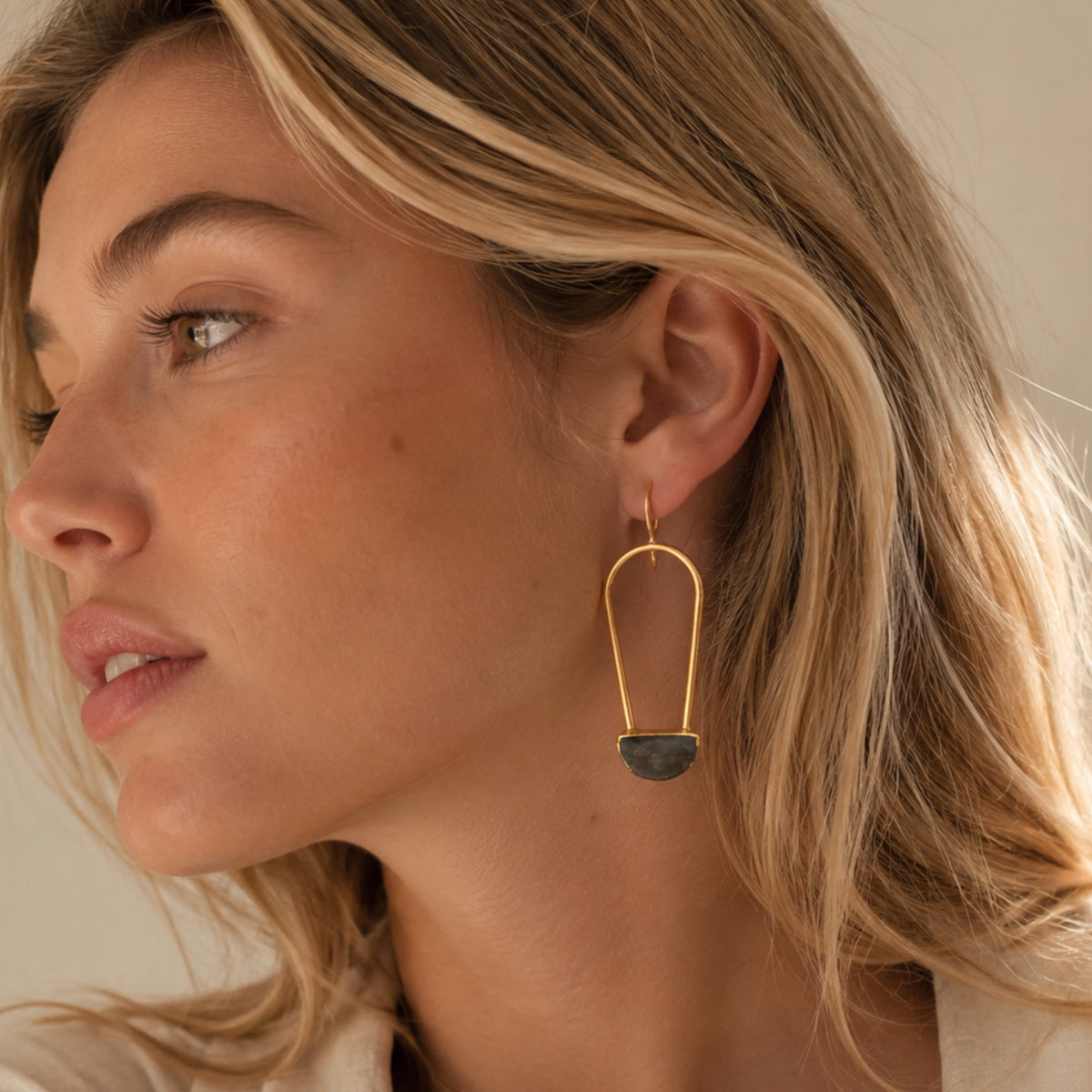 Blonde woman close up lifestyle image wearing gold plated abstract drop earrings with a faceted semi-circle labradorite center 