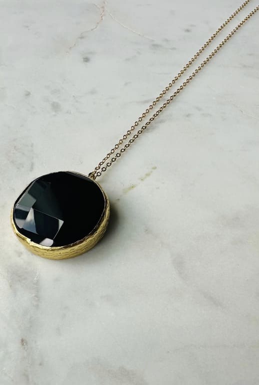 Close-up diagonal view of artisanal sustainable gold-plated round onyx pendant Necklace with a long chain on a marble surface