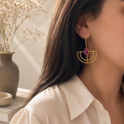 A close-up lifestyle shot of a woman wearing a gold geometric shaped dangle earring with a ruby teardrop gemstone, set against a soft, warm background with a vase of dried flowers.