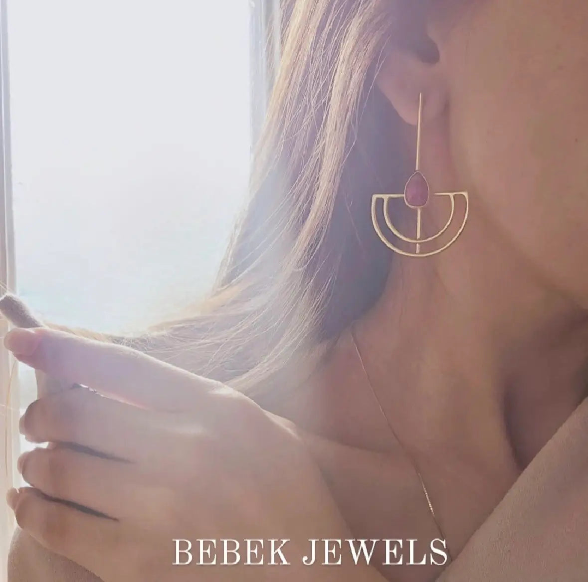Close-up of a woman wearing gold-plated geometric ruby earrings with 'BEBEK JEWELS' branding