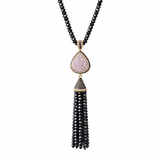 A sapphire beaded necklace featuring a teardrop-shaped light purple druzy stone pendant with gold-tone detailing, a small black beaded cap, and a long bead tassel