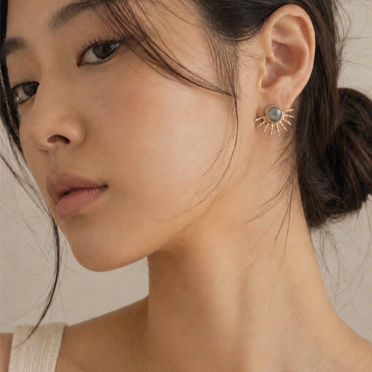 Close-up of a woman wearing a gold sunburst stud earring with a round gray-blue stone, shown against her ear and neck.