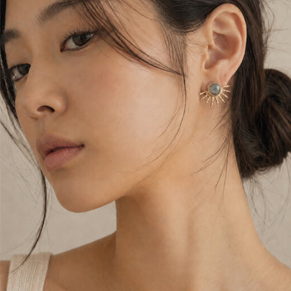 Close-up of a woman wearing a gold sunburst stud earring with a round gray-blue stone, shown against her ear and neck.