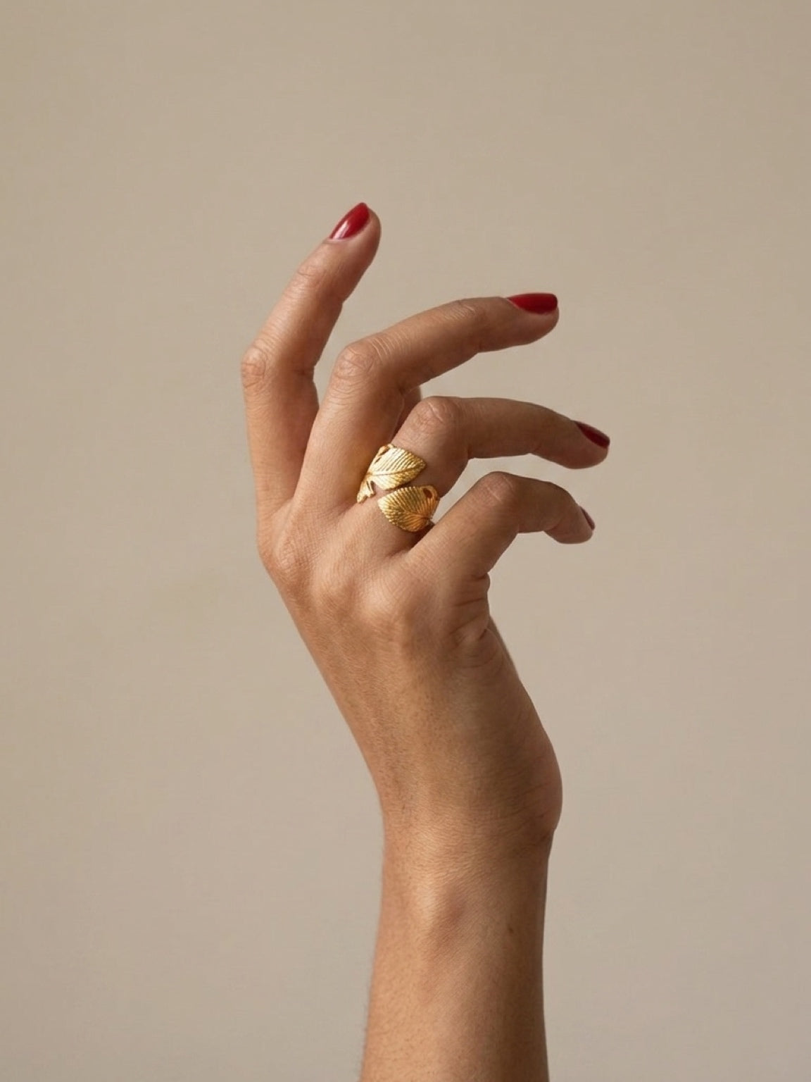 A close-up of a hand with red nail polish wearing a gold feather ring, set against a solid beige background.