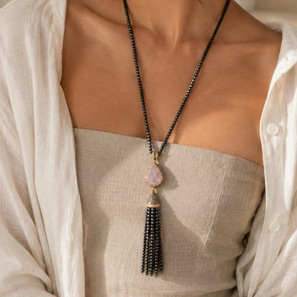 A close-up of a woman wearing a long sapphire beaded necklace with a teardrop-shaped light purple geode pendant and a dangling bead tassel, styled over a beige top and white shirt.