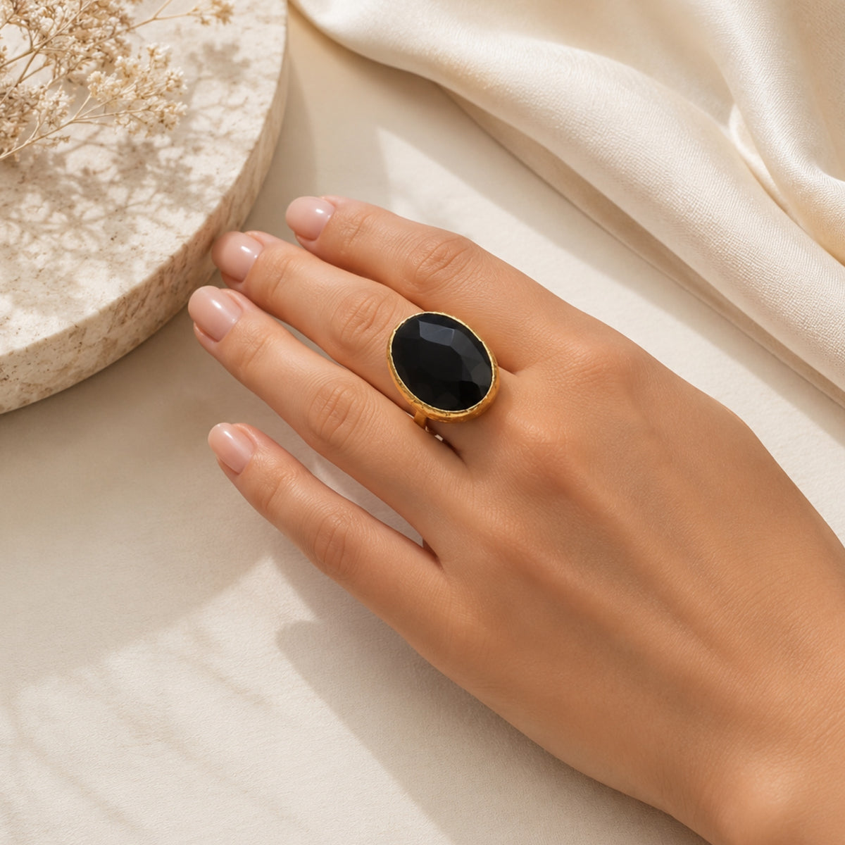 A hand wearing a handmade gold-plated large faceted oval onyx ring with against a neutral  setting