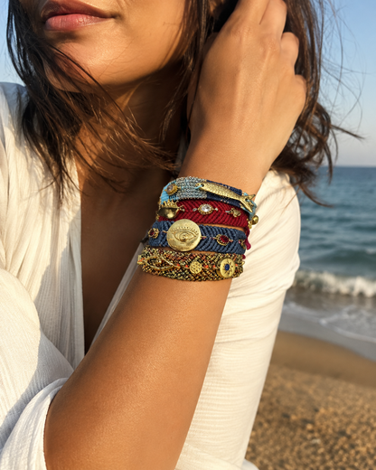 A close-up of a woman's wrist at the beach wearing a stack of colorful woven bracelets with gold-toned coin details and crystals 