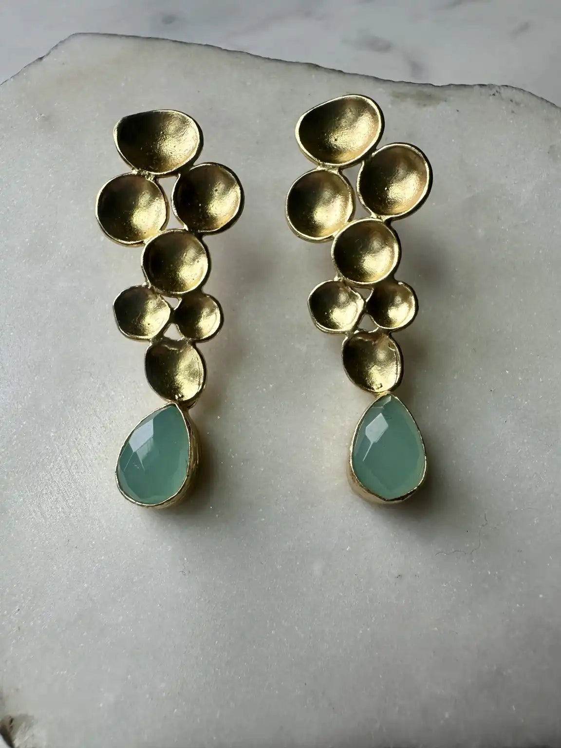 A pair of gold-plated contemporary drop earrings with a cluster of textured gold discs and a faceted aqua chalcedony teardrop gemstone at the bottom, displayed on a white stone surface.
