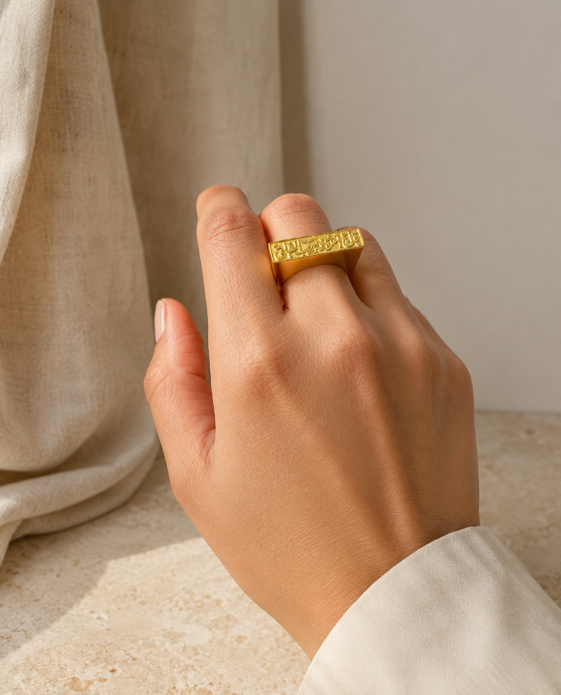 Hand wearing A gold plated bar ring with Arabic calligraphy engraved on it displayed on a neutral surface