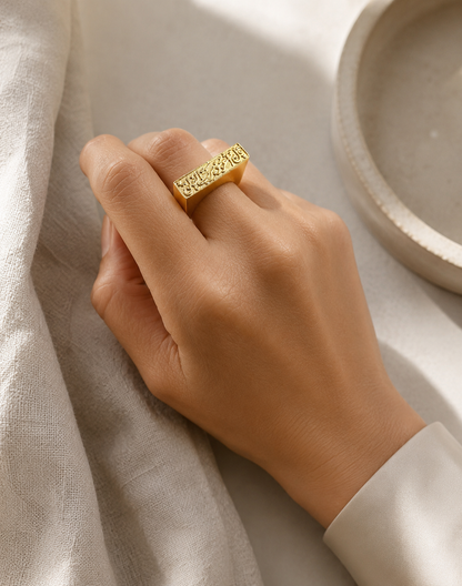 A Hand wearing A gold plated bar ring with Arabic calligraphy engraved on it displayed on a neutral surface