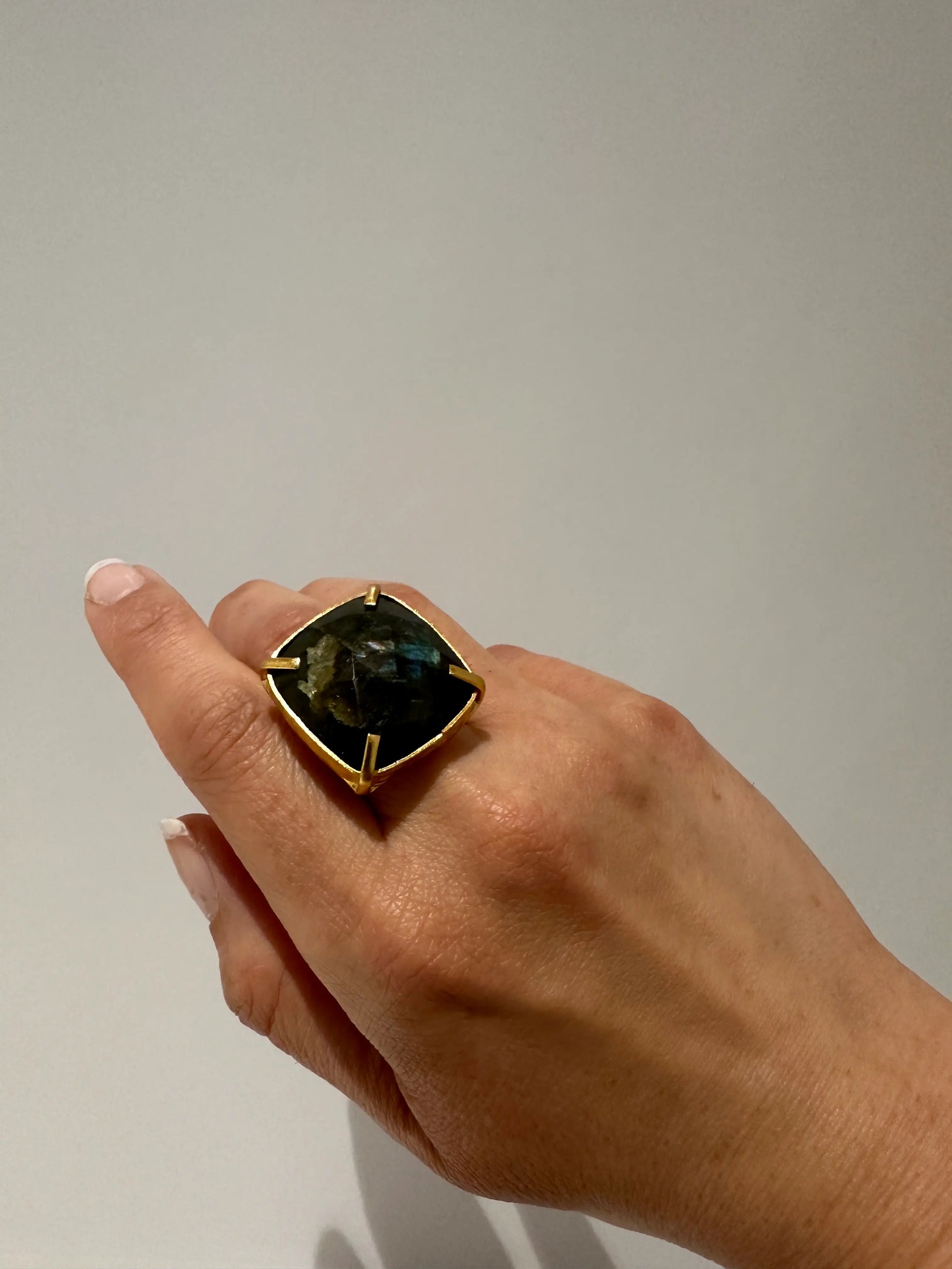 Hand wearing a ring with a square labradorite gemstone on a plain background