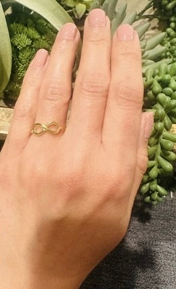 Hand wearing adjustable gold-planted infinity ring with an infinity symbol against a green plant background