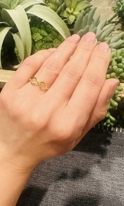 Hand wearing a gold-plated infinity ring with plants in the background