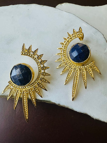 Gold sun-shaped earrings with blue gemstones on a white shell background