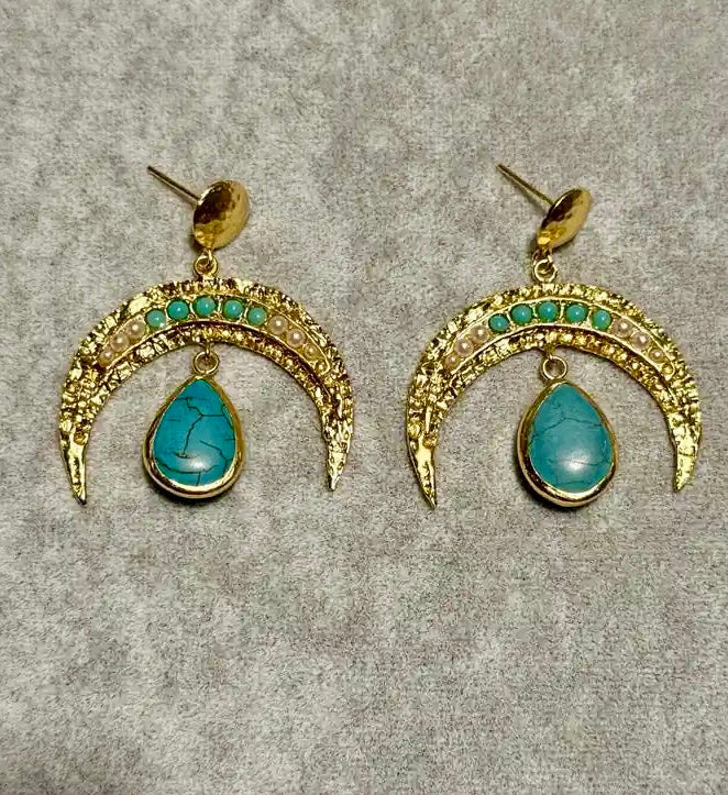A pair of handmade gold-plated crescent-shaped drop earrings with small turquoise and pearl gemstones along the top arch and a larger teardrop turquoise stone hanging below, displayed on a textured gray background.
