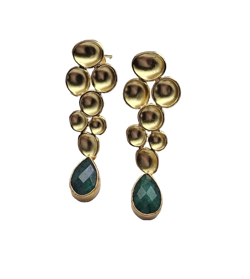 A pair of gold drop earrings featuring  geometric gold metal design and a faceted green teardrop emerald gemstone at the bottom, set against a plain white background.