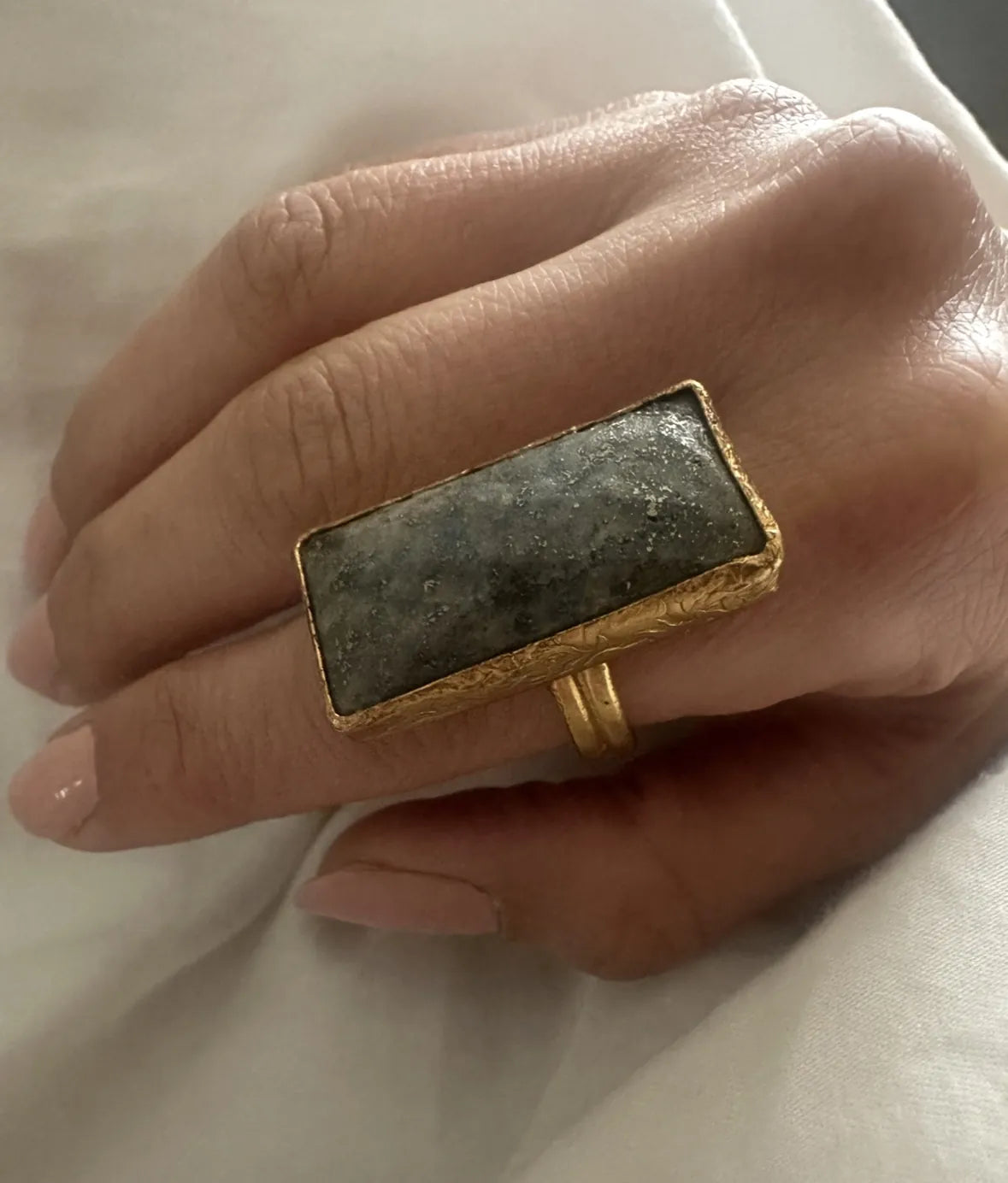 Gold cocktail ring with a rectangular dark gray gemstone on a hand against a white background
