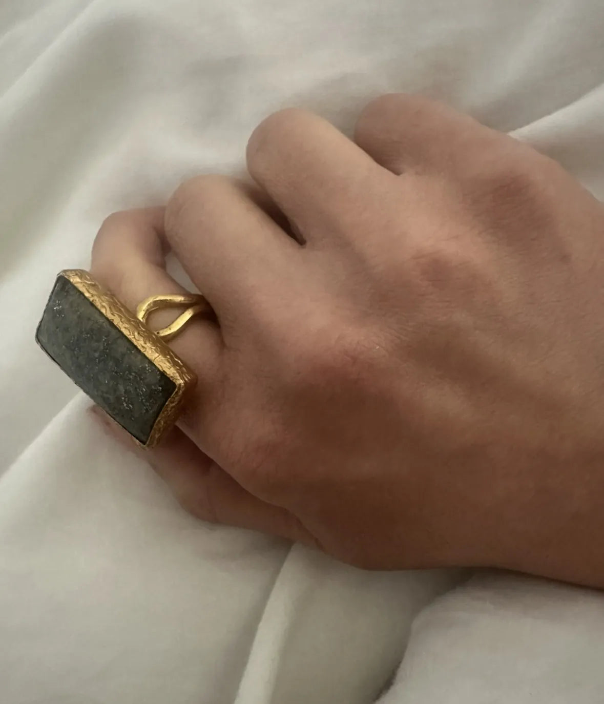 Hand wearing a gold ring with a dark gray lapis lazuli stone on a white fabric background