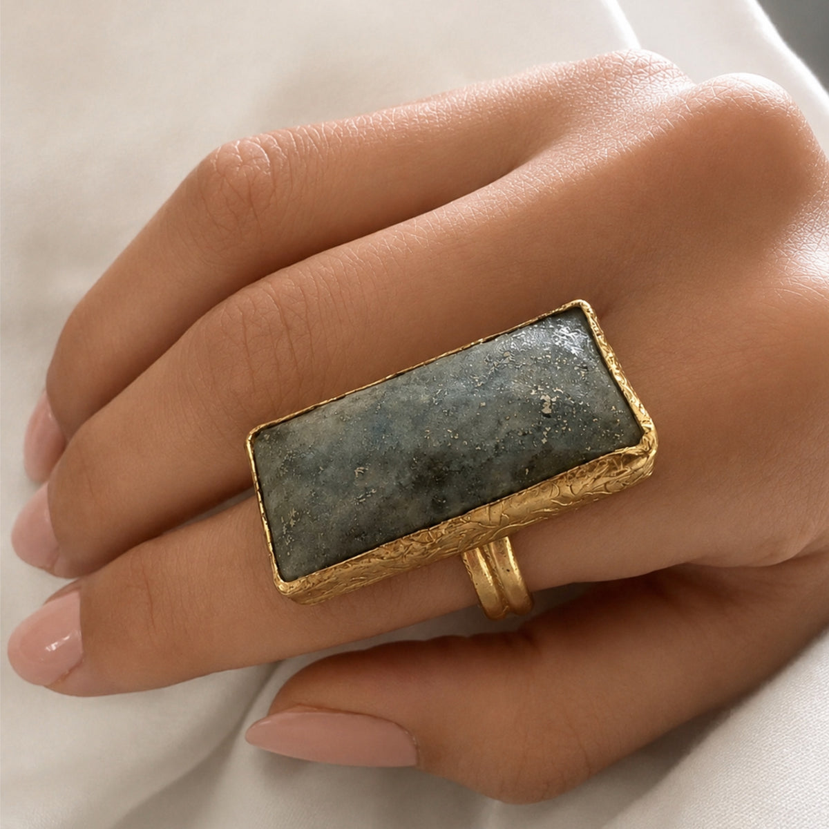 Hand wearing a gold ring with dark gray lapis lazuli stone on a white fabric background