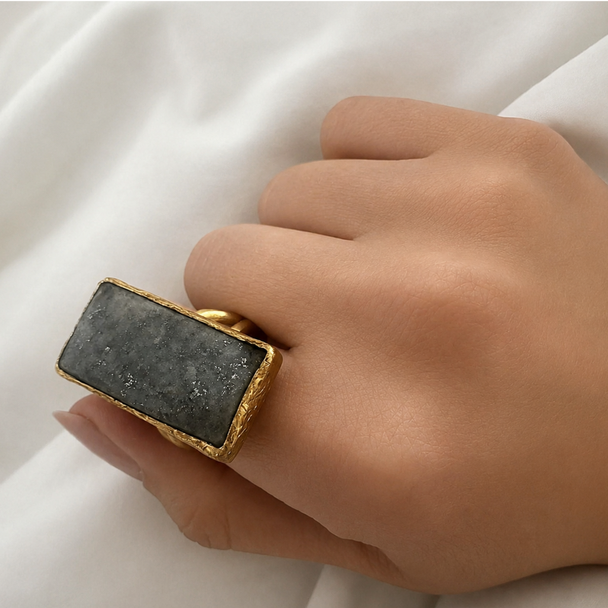 Hand wearing gold ring with a dark gray lapis lazuli stone on a white fabric background