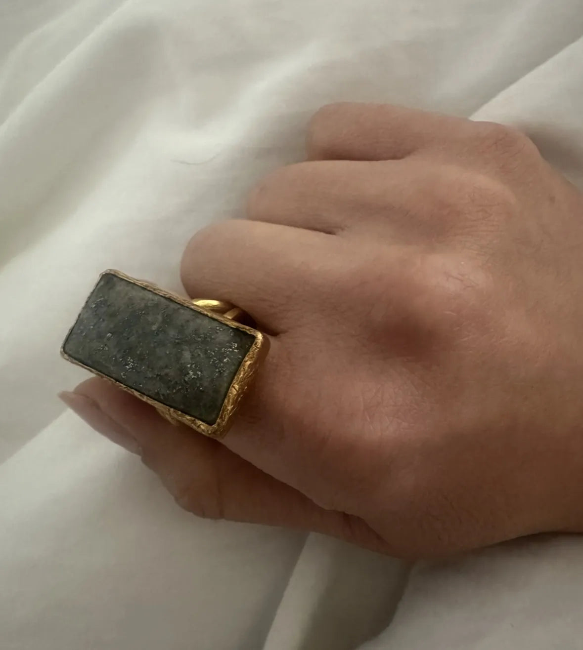 Hand wearing a goldring with a rectangular gray lapis lazuli stone on a white fabric background