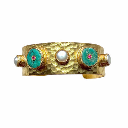 A handmade hammered gold cuff bracelet featuring turquoise inlay circles with small red zircon centers and white pearls shown on a plain white background