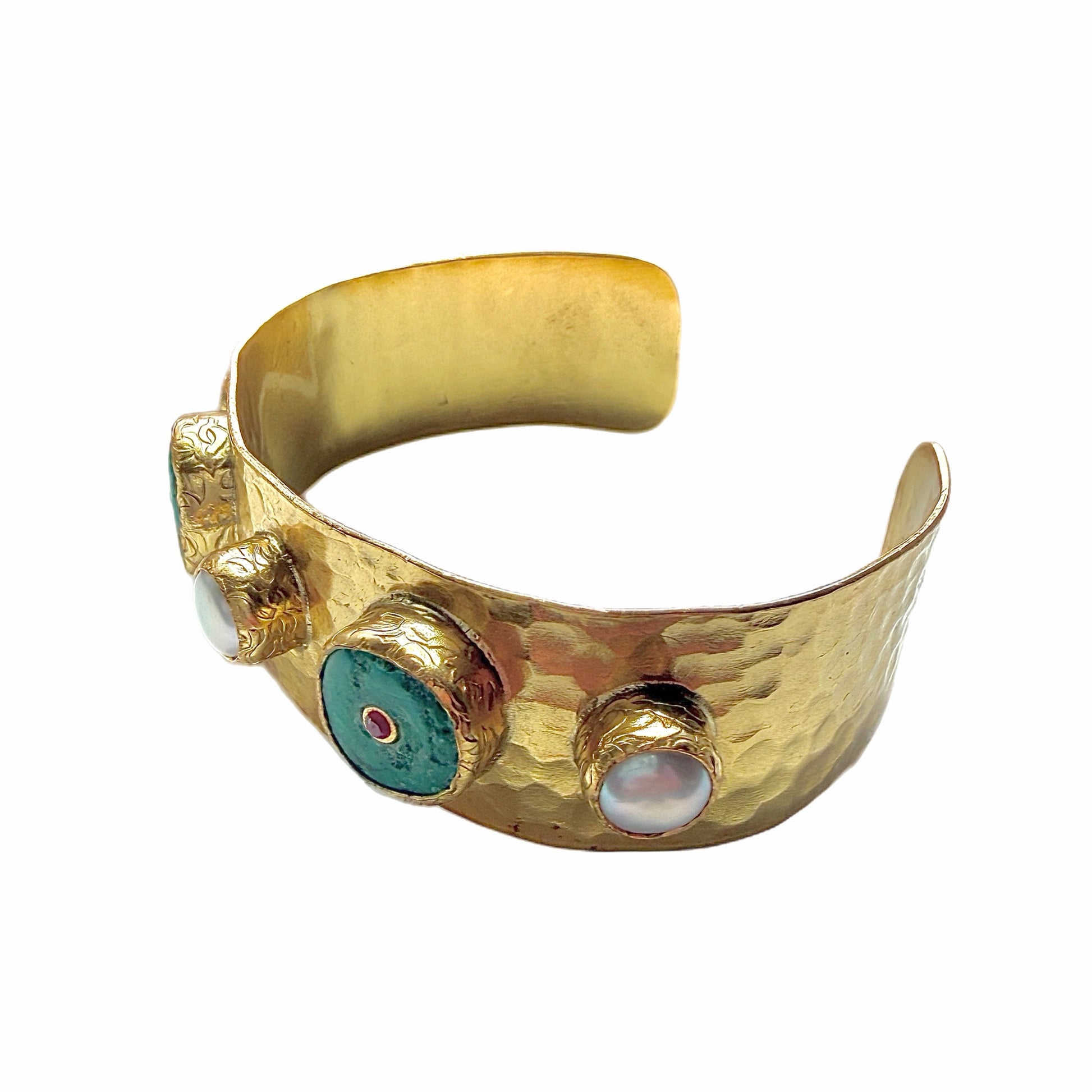 A handmade hammered gold cuff bracelet featuring turquoise inlay circles with small red zircon centers and white pearls shown on a white backdrop